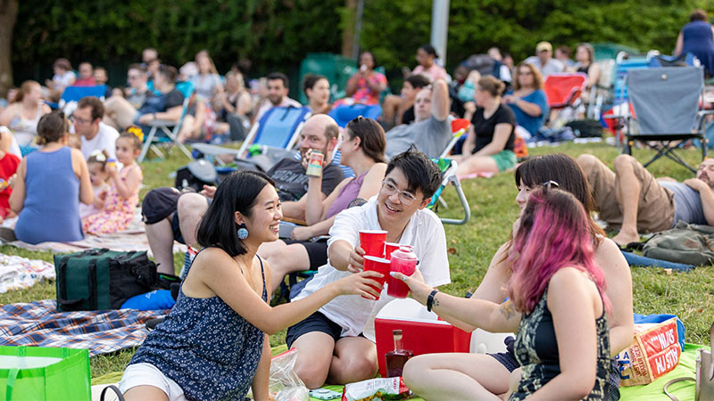 Young Friends members raising their glasses while having a picnic at an outdoors concert.